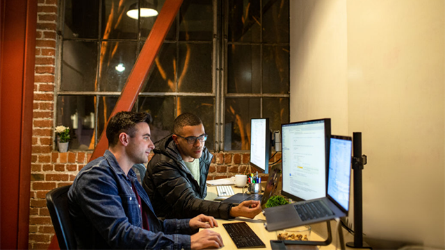 Two people sit at desks in consultation while facing computers.
